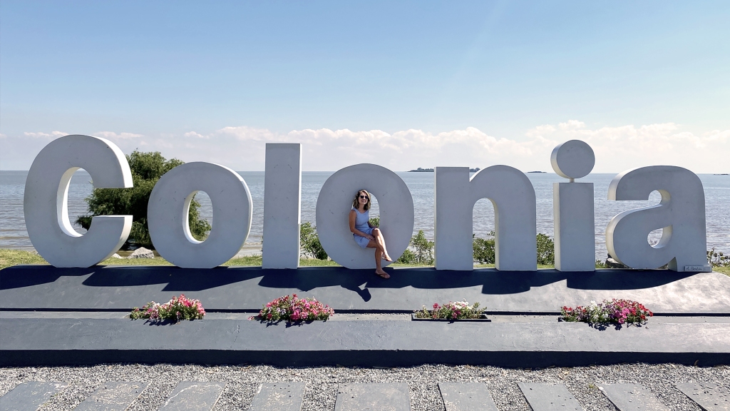 A large, white letter sign reading the name of the town "Colonia" in front of a river walk in Uruguay.