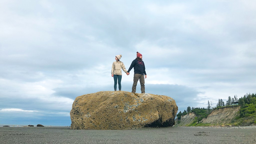A young man holding hands and standing on top of a large rock on a beach in Homer, Alaska.