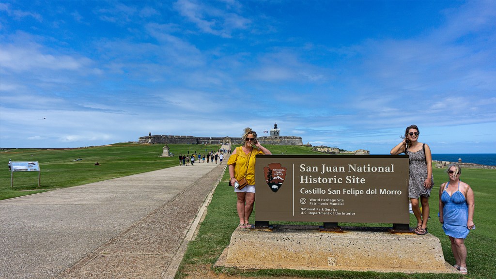 Three woman standing in front of the San Juan National Historic Site Castillo San Felipe del Morro sign in Puerto Rico. In the distance you can see the fort.