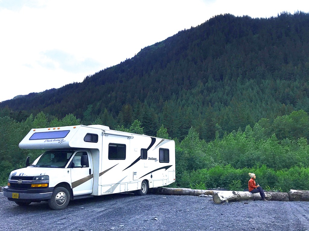 A large RV parked in front of a tree covered mountain, with a young woman sitting on a stump outside of the RV in Alaska.