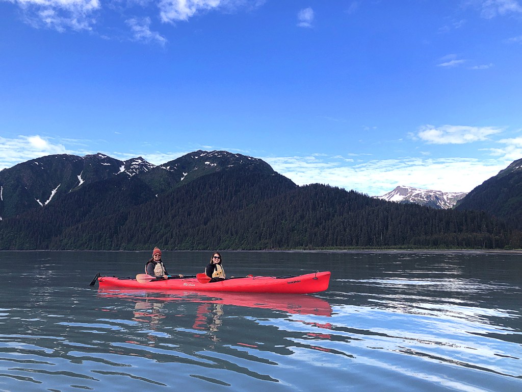A young man and woman inside of a red tandem kayak in a bay surrounded in mountains, in Seward, Alaska.