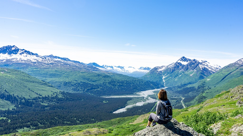 A woman sitting on the edge of an overlook of a green valley with a river below and a glacier in the distance in Alaska.