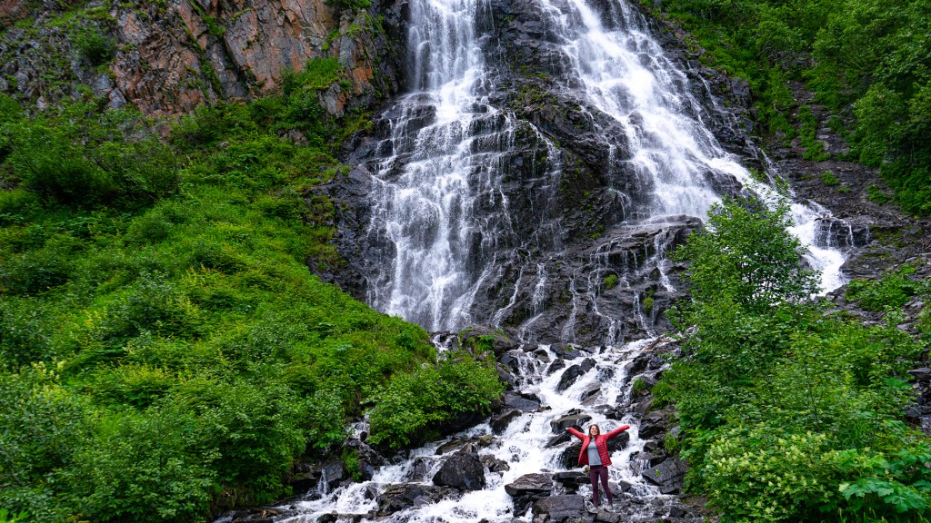 A young woman standing in front of a tall waterfall in Valdez, Alaska.