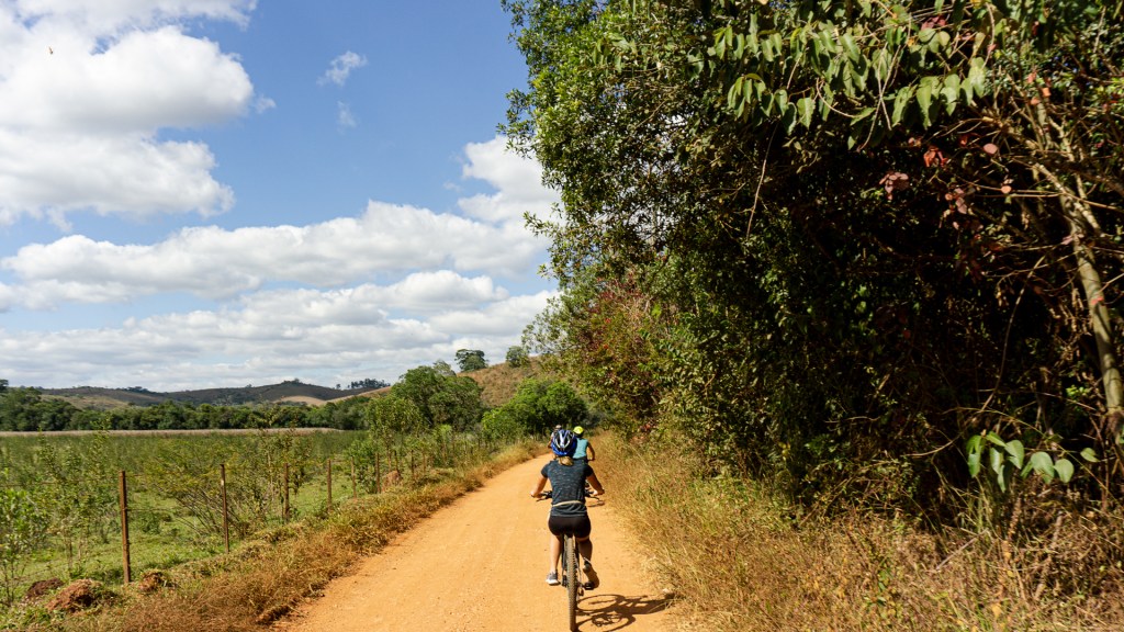 Two people riding bicycles down a dirt road with helmets on. There is a field on one side and trees on the other side of the dirt road. Bichinho, Tiradentes