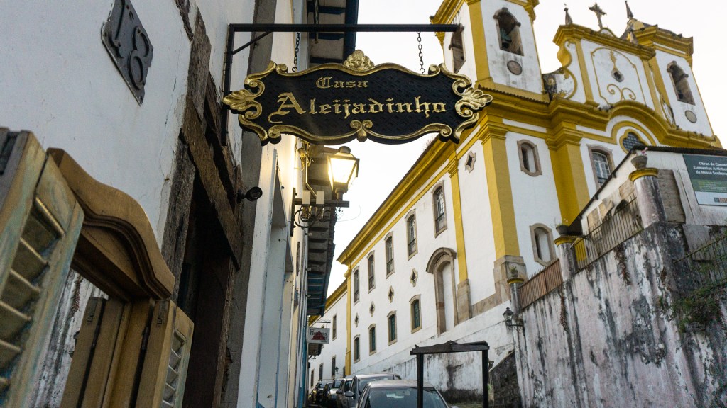 A gold plated sign reading Casa Aleijadinho, with a large white and yellow 18th century church in the background.