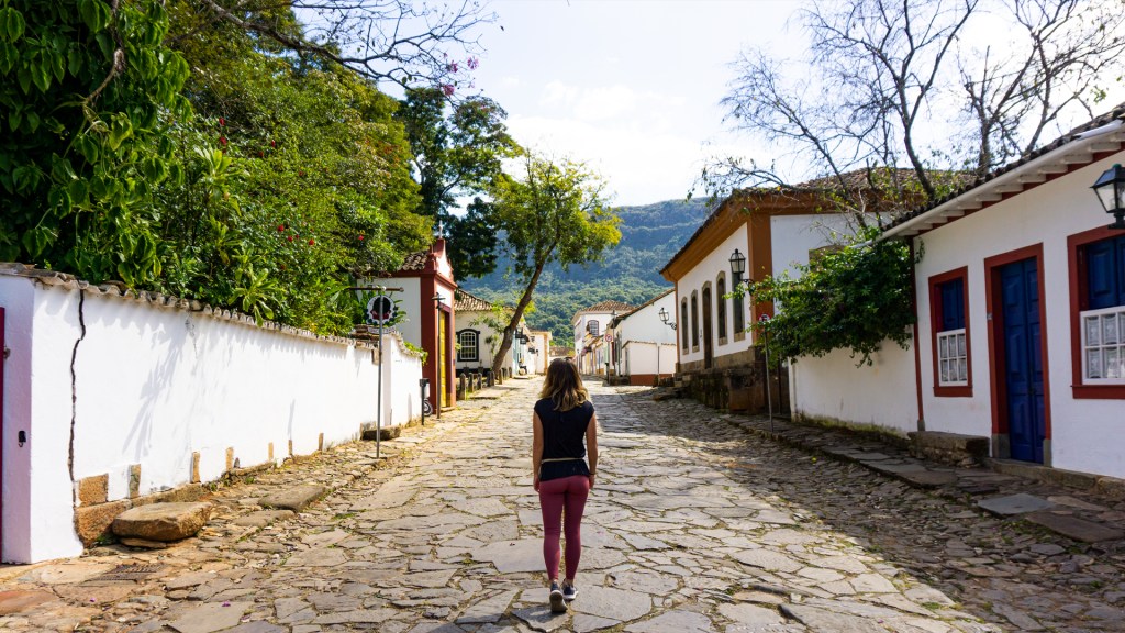 A woman walking down a cobblestone street lined with colorful colonial buildings and green trees and bushes, with mountains in the distance. Tiradentes, Minas Gerais, Brazil.