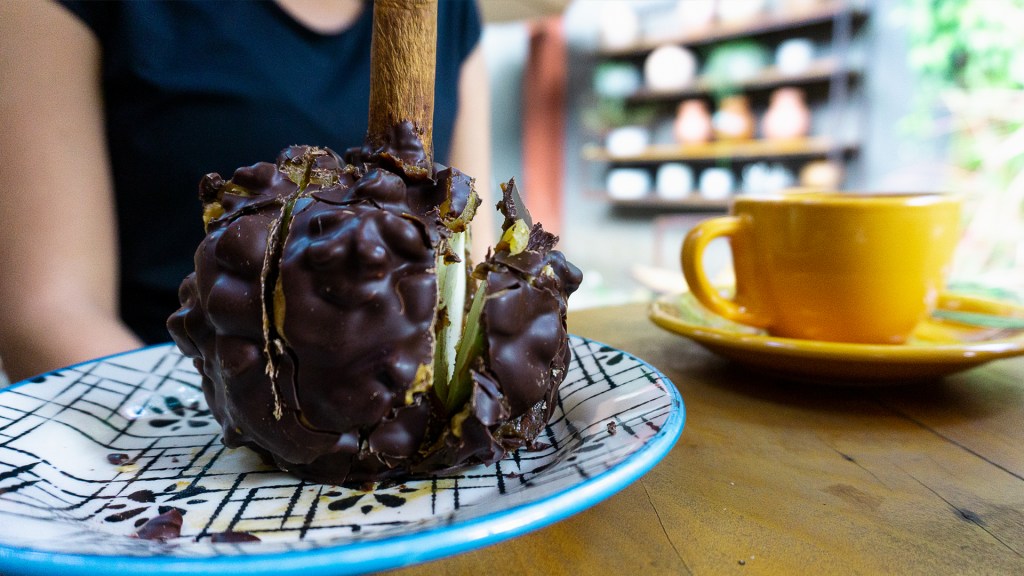 A gourmet caramel apple covered in chocolate and nuts, sitting on a plate next to a coffee cup in a cafe. Tiradentes, Minas Gerais, Brazil.