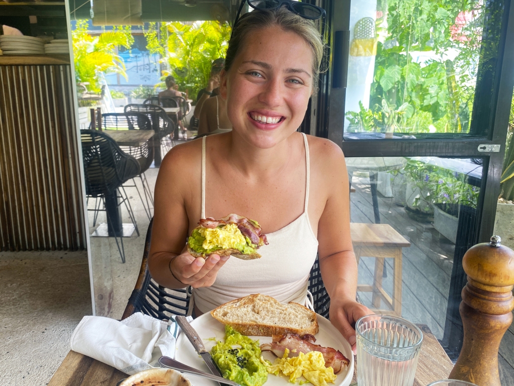 A woman at the Avocado Factory Cafe in Canggu, Bali eating a slice of avocado toast topped with scrambled eggs and bacon.
