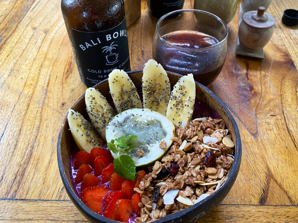 A smoothie bowl topped with berries, granola, and slices of banana inside of a coconut bowl. There is a glass of cold brew coffee from Bali Bowls, Canggu, Bali.