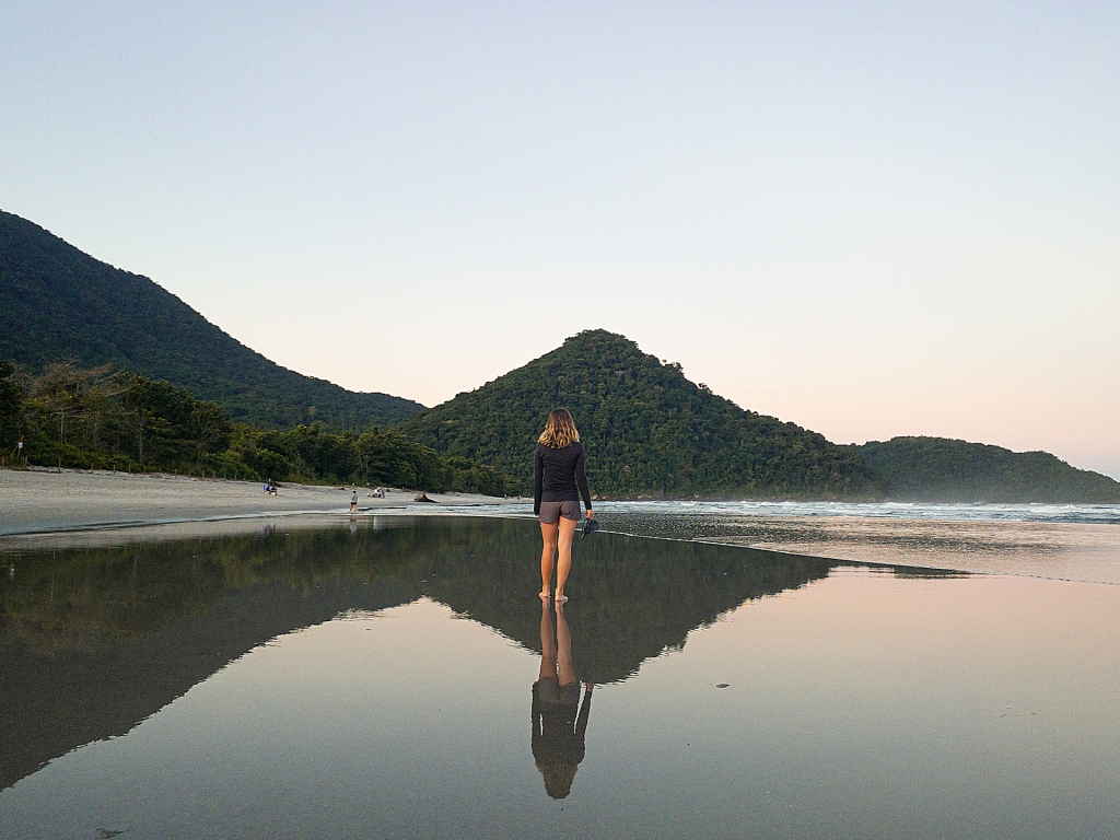 A woman walking on the beach in Ubatuba, Sao Paulo, Brazil. There is a mountain in the backdrop and a reflection below.