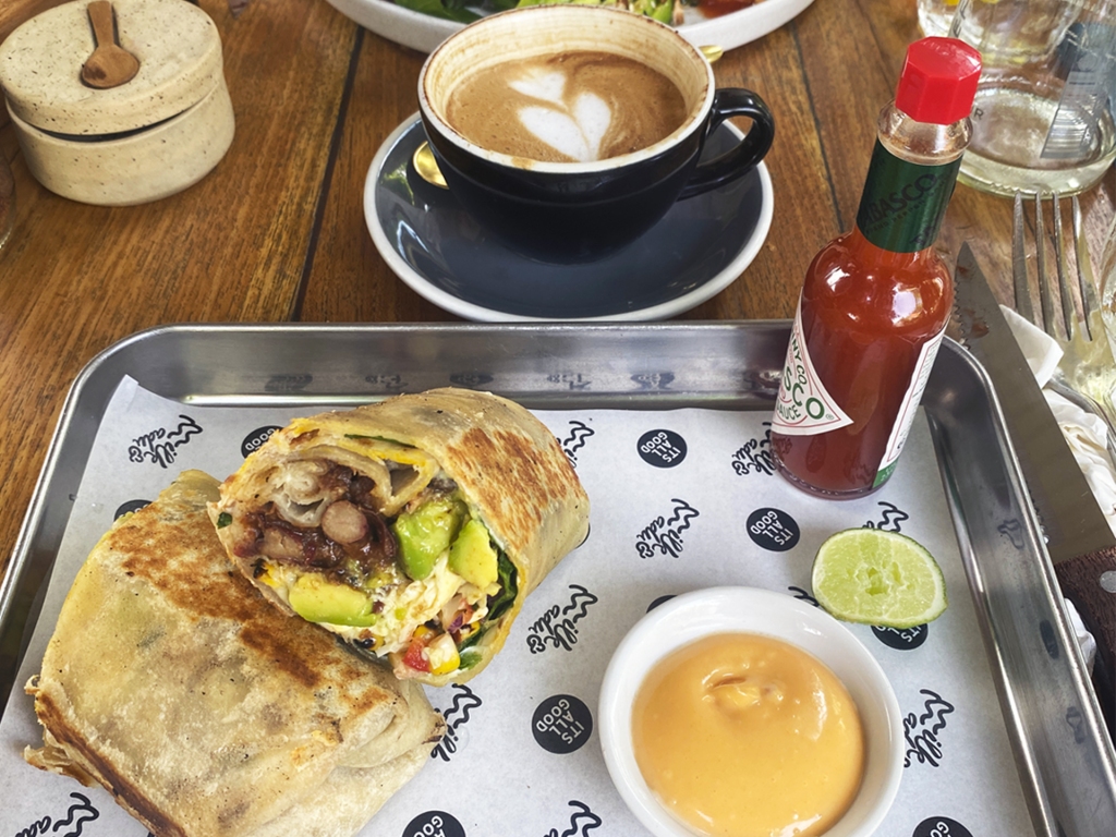 A tray of food holding a breakfast burrito filled with eggs, avocado, sausage, and cheese. A cappuccino at milk and madu cafe in Canggu, Bali.