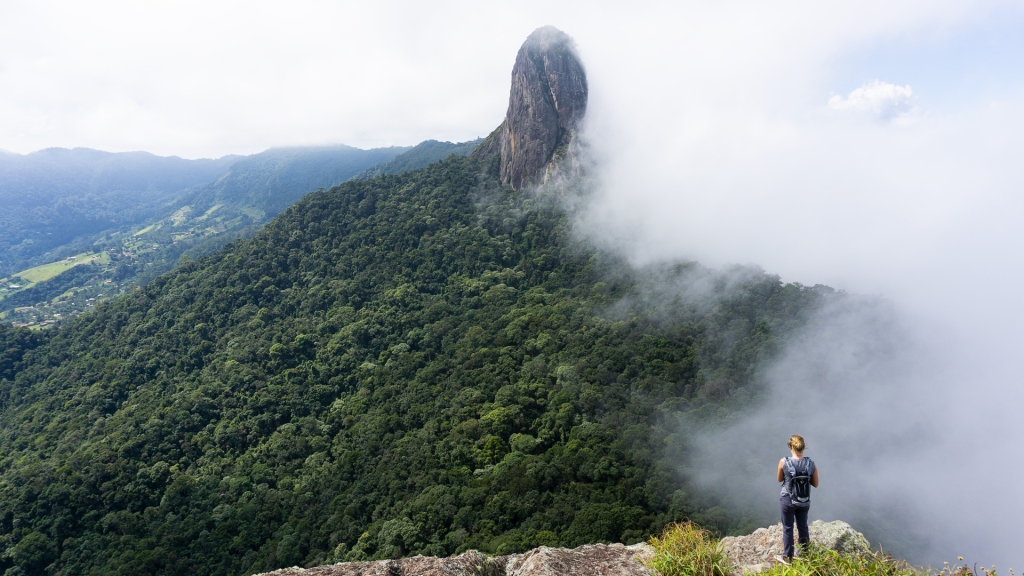 The Pedra do Baú via the Ana Chata Trail in São Bento do Sapucaí, Sao Paulo, Brazil. A woman looking at a giant rock formation from atop a mountain in the Mantiqueira Mountains.