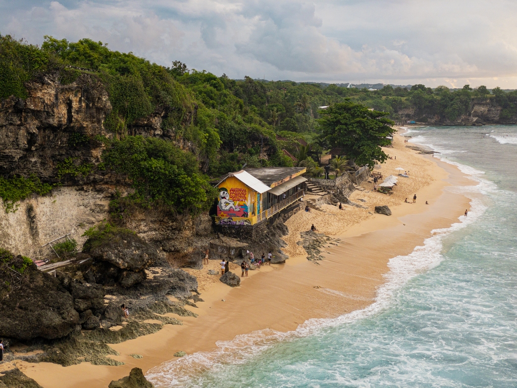 An aerial photograph of Balangan Beach in Uluwatu, Bali. There is a cliff next to the beach and a yellow hut with graffiti on the side.