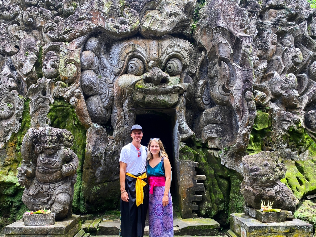 A young man and woman standing in front of a cave temple door that is decoratively carved into the rock face opening to look like a face.