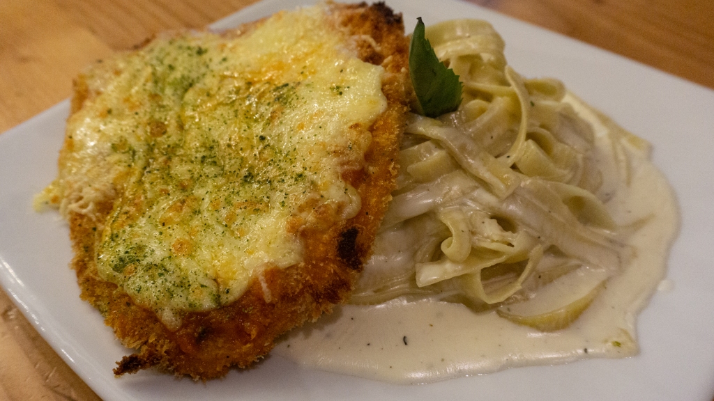 A piece of breaded chicken covered in cheese, sitting on top of a pile of alfredo pasta.