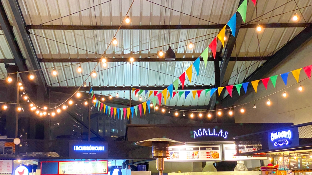 A market full of different restaurants with string lights and colorful flags strung from the ceiling.