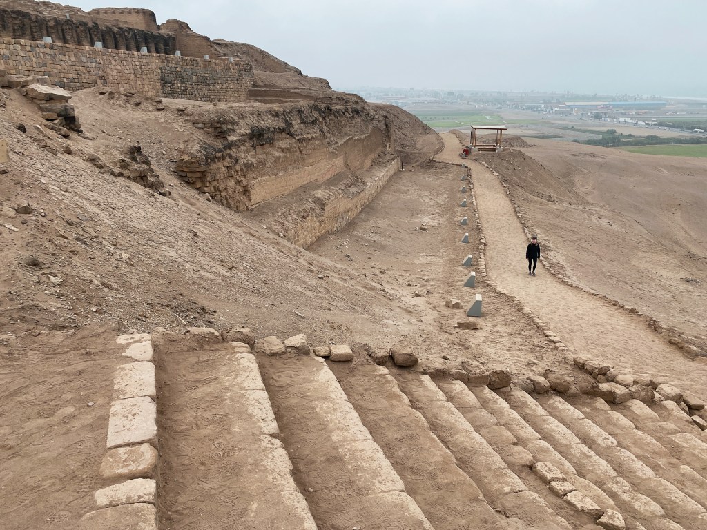 A woman walking down a dirt path in front of an ancient Peruvian pyramid called the Pachacamac Ruins.