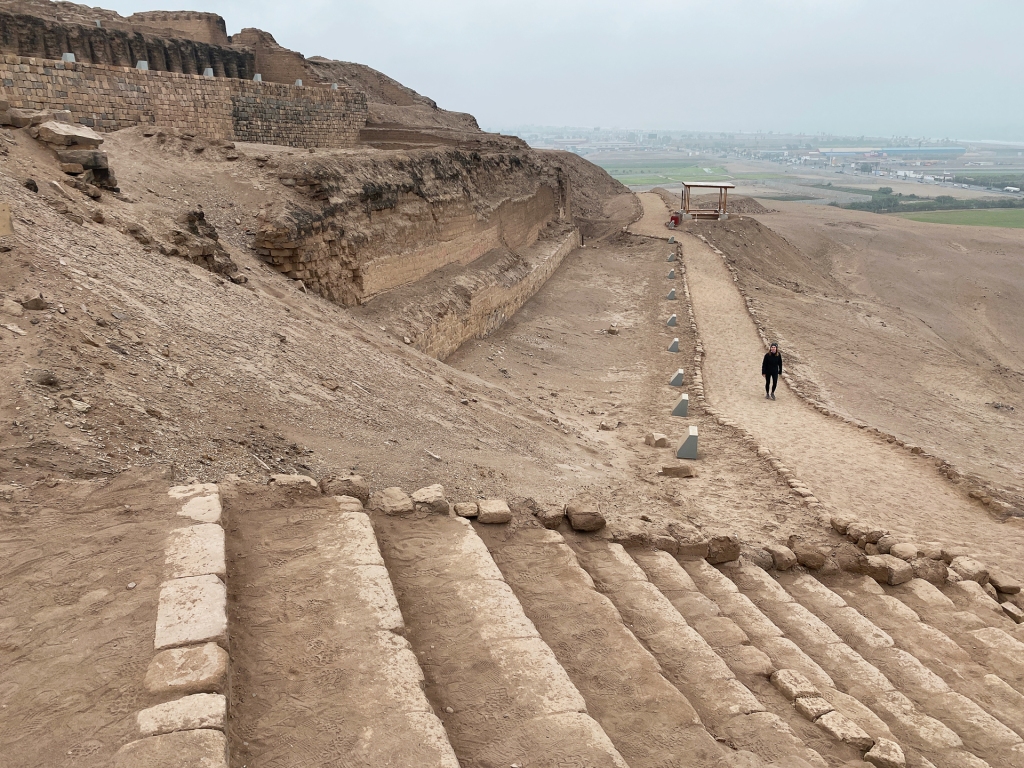 A woman walking down a dirt path in front of an ancient Peruvian pyramid called the Pachacamac Ruins.