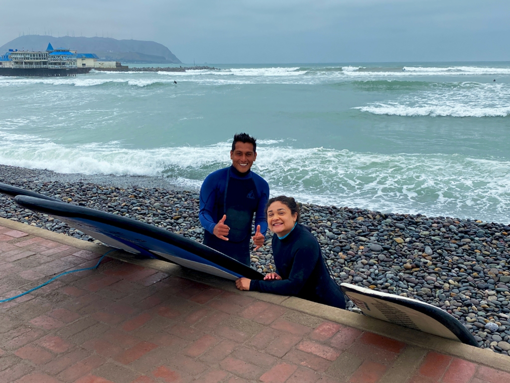 A man and a woman wearing wetsuits, holding surf boards, in front of the ocean in Lima, Peru.