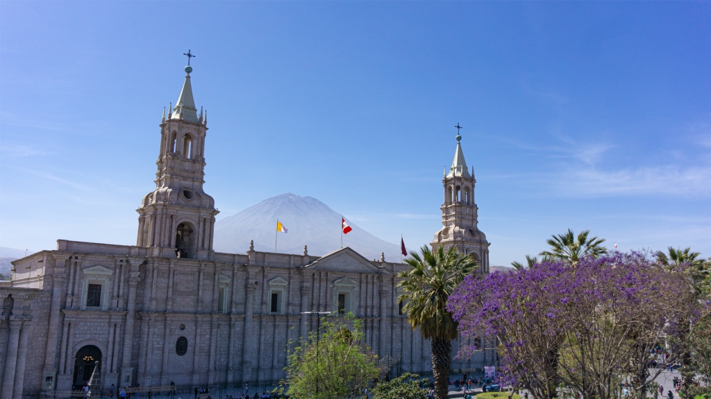 A large, white cathedral with an impressive volcano between it's two steeples in the Plaza de Armas of Arequipa, Peru.