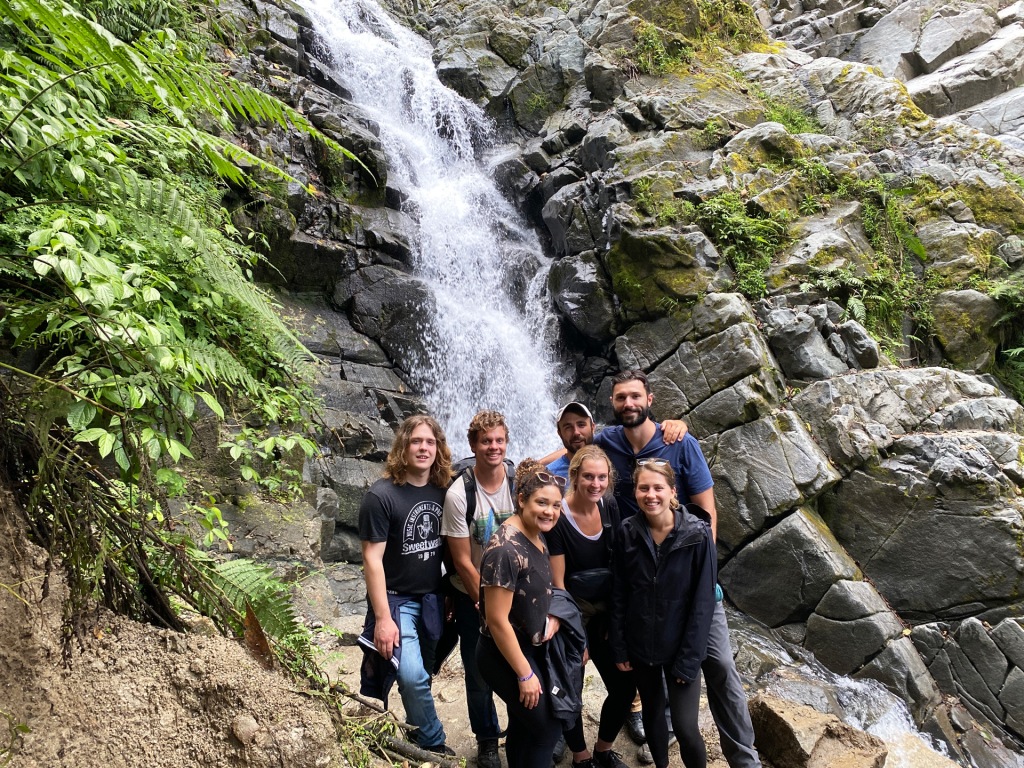 A group of seven young adults standing in front of a waterfall in Aguas Calientes, Peru.