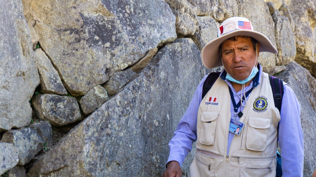 An older man who is a guide for the Machu Picchu ruins, standing in front of one of the ancient walls within the ruins.