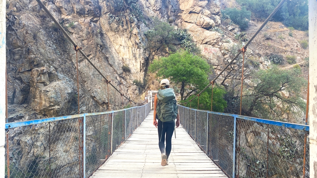 A woman wearing a green backpack, walking across a wooden bridge in Colca Canyon, Peru.