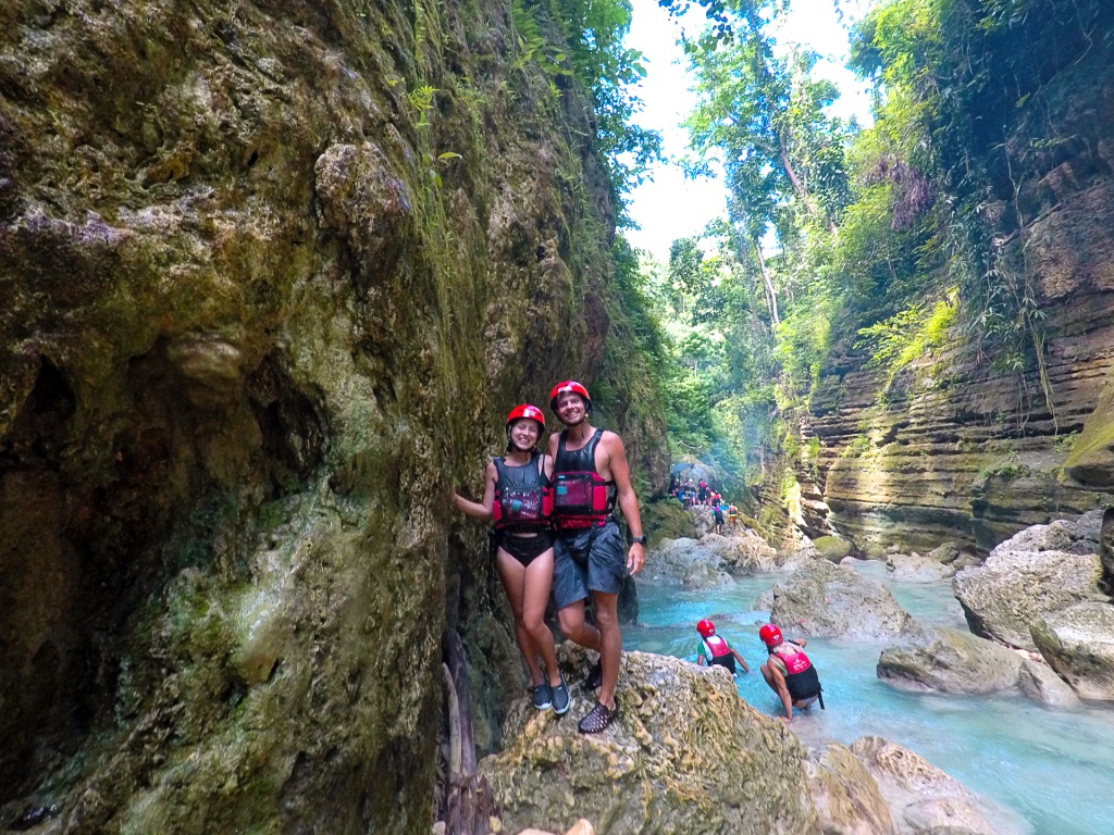A man and a woman wearing life jackets and red helmets, standing in a canyon with a very blue river.