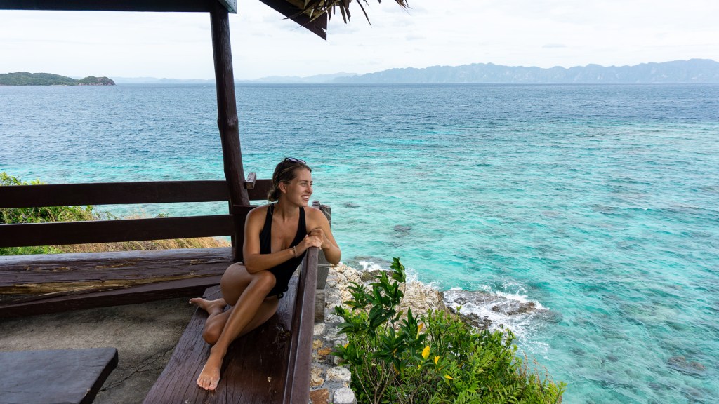 A woman sitting on a wooden bench and looking at the bright blue water of the ocean in Coron, Palawan, Philippines.