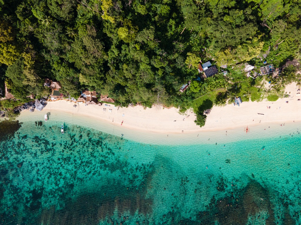 An aerial photo of a bright blue beach lined with green trees in Palawan, Philippines.