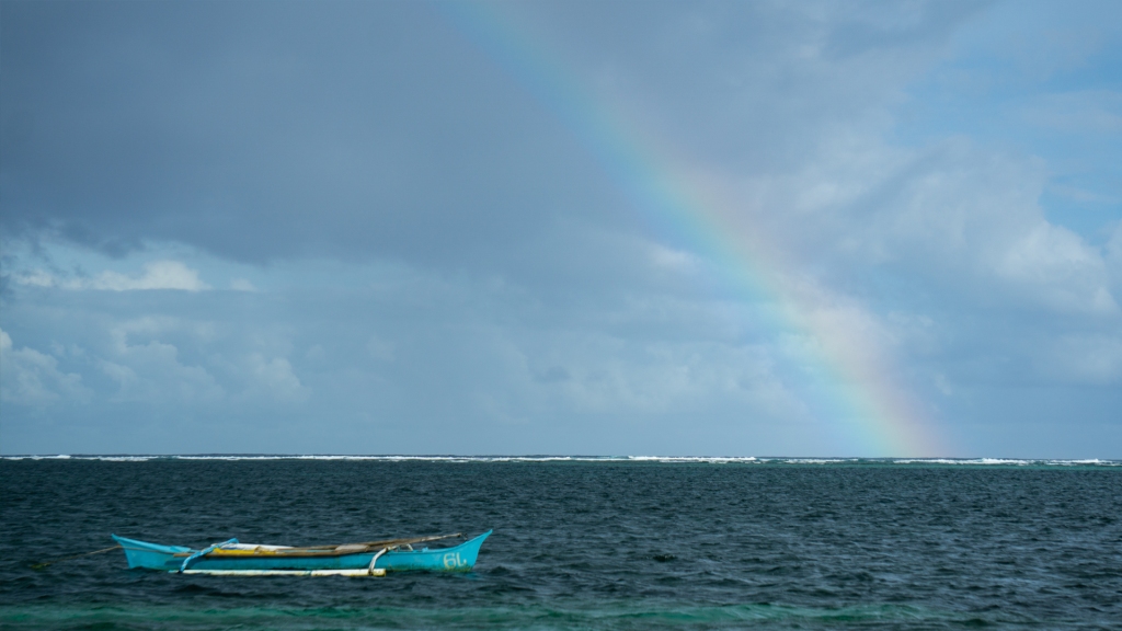 A blue wooden boat in the ocean, with a rainbow in the sky in Siargao, Philippines.